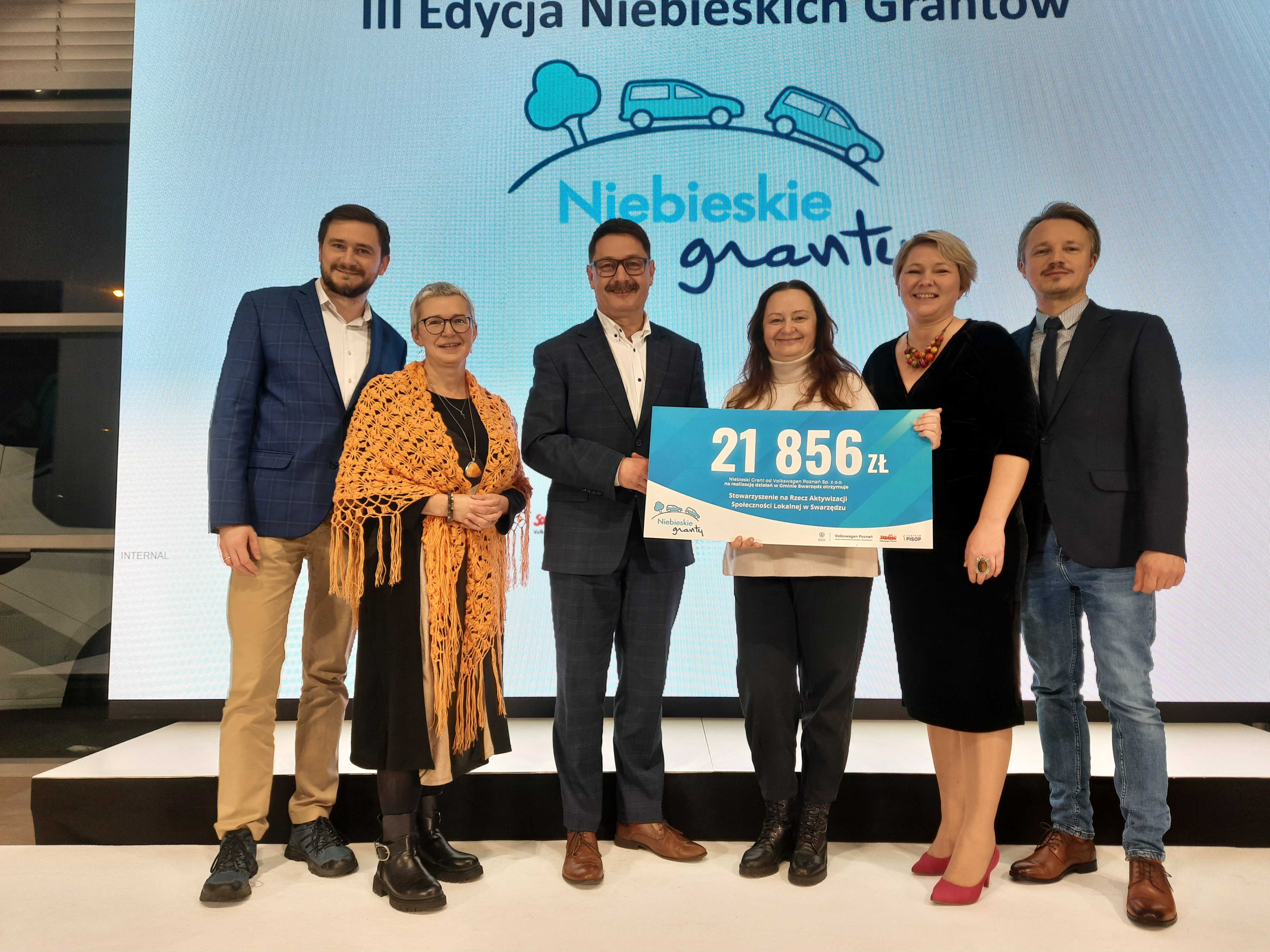 Wręczenie czeku 21 856 zł w ramach Niebieskich Grantów — grupa osób na scenie, para w centrum trzyma duży czek.