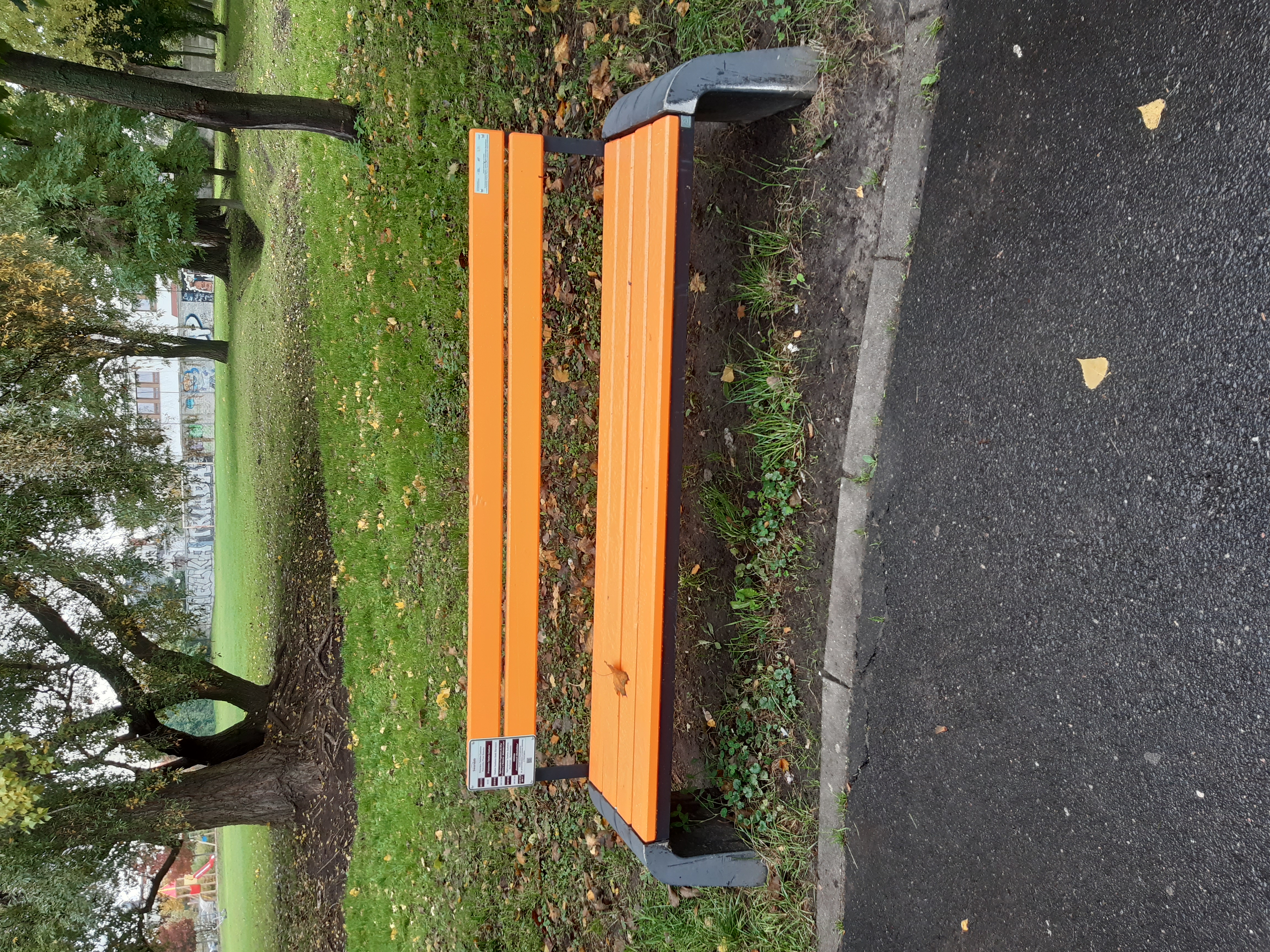 Pomarańczowa ławka w parku przy ul. Pogodnej w Swarzędzu; z boku tabliczka z numerami telefonów pomocowych.