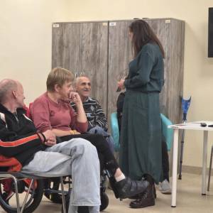 Rozmowa w trakcie zajęć; prowadząca stoi przy uczestnikach, obok wózek i kule, sala ŚDS.