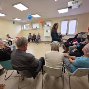 Zajęcia grupowe w jasnej sali; prowadząca mówi do uczestników siedzących w półokręgu.