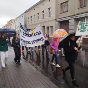 Pochód w deszczu. Uczestnicy z parasolami niosą transparent „Zdrowie psychiczne to nasza wspólna sprawa” i tablicę „Bądź dla siebie dobry”.