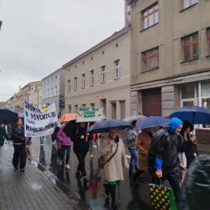 Kolejne ujęcie pochodu w deszczu. Uczestnicy z parasolami niosą transparent o zdrowiu psychicznym oraz tablicę „Bądź dla siebie dobry”.