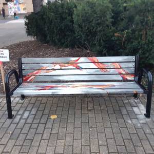 Ławka z artystycznym malunkiem w odcieniach czerwieni i szarości, z tabliczką ‘świeżo malowane’ obok.
