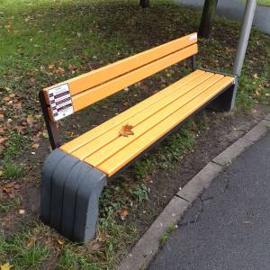 Żółta ławka przy ścieżce w parku, z tabliczką projektu na oparciu i liściem leżącym na siedzisku.