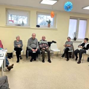 Grupa seniorów siedzi w kręgu na zajęciach w jasnej sali, rozmawiają i uczestniczą w spotkaniu integracyjnym