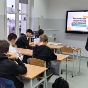 Uczniowie pracują przy ławkach, a prowadząca stoi obok; na ekranie widoczny jest slajd ‘Budowanie odporności psychicznej’