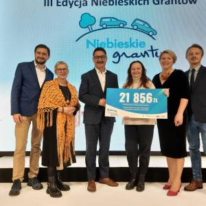 Wręczenie czeku 21 856 zł w ramach Niebieskich Grantów — grupa osób na scenie, para w centrum trzyma duży czek.