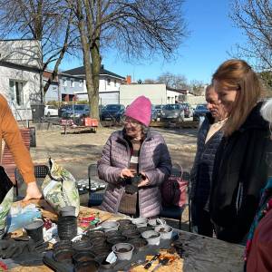 Grupa seniorów i opiekunów uczestniczy w zajęciach ogrodniczych na świeżym powietrzu przy stole z sadzonkami.