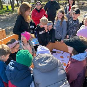 Dzieci i opiekunowie stoją wokół drewnianej skrzyni podczas wspólnych zajęć ogrodniczych.