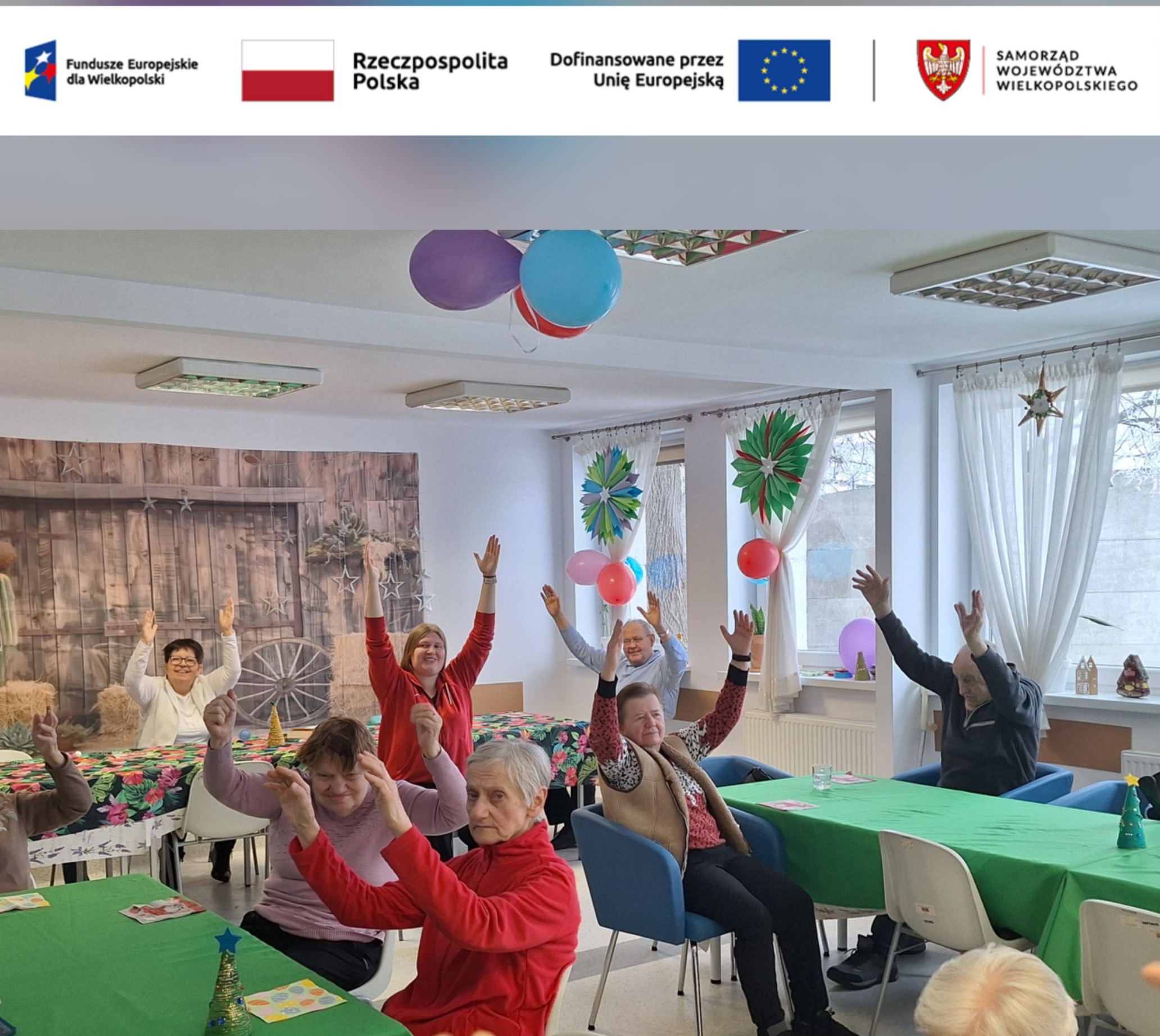 Seniorzy siedzą przy stolikach i wspólnie unoszą ręce podczas zajęć gimnastycznych w sali Dziennego Domu Pomocy, ćwiczenia grupowe