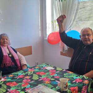 Dwoje seniorów siedzi przy stole i ćwiczy ręce, podnosząc lekkie hantle podczas zajęć z rehabilitantką w DDP.