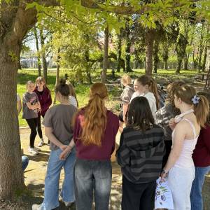 Grupa młodzieży stoi w cieniu drzew i rozmawia podczas warsztatów w ogrodzie społecznym
