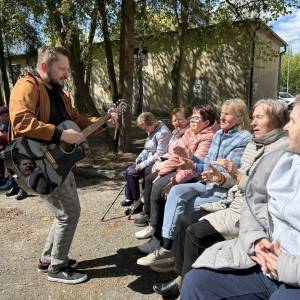 Mężczyzna gra na gitarze dla grupy seniorów siedzących na zewnątrz podczas spotkania w ogrodzie społecznym