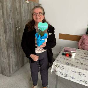 Seniorka stoi w sali i trzyma ozdobę z niebieskim sercem oraz małym bukietem kwiatów