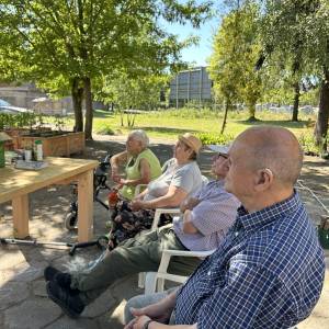 Cztery osoby wysłuchują wykładu na ogrodzie Centrum Usług Społecznych w Swarzędzu. W tle widać trawiastą polanę i drzewa w ogrodzie.