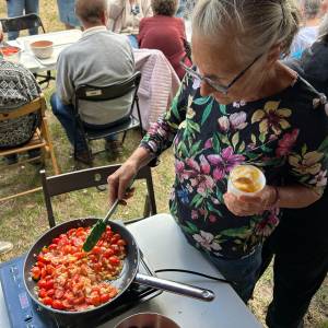 Jedna z seniorek szpatułką silikonową miesza pomidory z cebulką smażące się na patelni. W lewej ręce trzyma plastikowy kubek z koktajlem owocowym.