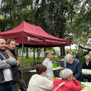 Seniorki i seniorzy pracują przy stołach plenerowych, szatkując kapustę i krojąc ogórki na warsztatach przetwórczych. Nad grupą rozstawiony jest bordowy namiot Centrum Usług Społecznych w Swarzędzu, obok którego stoją opiekunowie i uczestnicy obserwujący przebieg zajęć w ogrodzie. Spotkanie odbywa się na świeżym powietrzu wśród zielonych drzew.