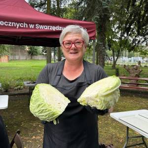 Pani Ania, instruktorka warsztatów przetwórczych, stoi w Ogrodzie Społecznym, trzymając w dłoniach w czarnych rękawiczkach dwie połówki dużej kapusty przygotowanej do kiszenia. Kobieta w okularach i czarnym fartuchu uśmiecha się do aparatu, a za nią widać czerwony namiot i parkową zieleń. Spotkanie odbywa się w ramach projektu 
