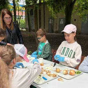 Dzieci z placówki 'Nasza Dziupla' wspólnie z opiekunkami obierają i kroją cebulę oraz warzywa przy stole w Ogrodzie Społecznym w Swarzędzu. Uczestnicy w rękawiczkach ochronnych pracują nad tacami ze składnikami w ramach międzypokoleniowych warsztatów przetwórczych. W tle widać zieleń ogrodu oraz sąsiadujący budynek.