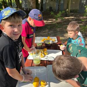 Grupa chłopców w rękawiczkach ochronnych wyciska i przygotowuje cytryny oraz limonki do napoju przy drewnianym stole plenerowym. Na tacach leżą połówki owoców, a obok stoją szklane dzbanki z wodą i miętą. Dzieci biorą udział w warsztatach kulinarnych zero waste w słonecznym Ogrodzie Społecznym.