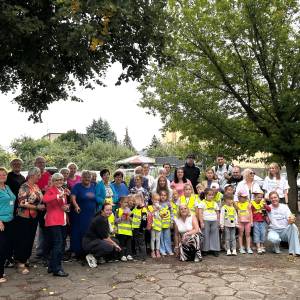 Pamiątkowe zdjęcie grupowe uczestników, instruktorów i organizatorów projektu „Ogród Pokoleń – siejemy smaki, zbieramy relacje” w Swarzędzu. Liczna, międzypokoleniowa grupa – seniorzy, dorośli oraz dzieci w żółtych kamizelkach odblaskowych – pozuje z uśmiechem na wybrukowanym placu pod rozłożystym drzewem Ogrodu Społecznego. W tle widoczna jest ogrodowa zieleń, zaparkowane samochody oraz zabudowania miejskie.